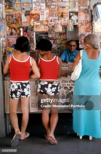 Couple wearing similar clothes buying magazines at a newsagent's kiosk. Rio de Janeiro, May 1977