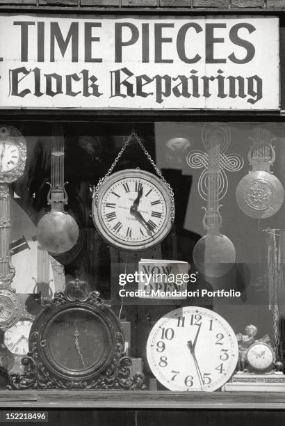 View of a watchmaker's shop window, whose sign 'Time pieces, clock repairing' is put among antique clocks and grandfather clocks. New York .