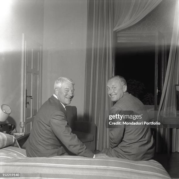 Jean Gabin and Bourvil are seated on a bed in a hotel room and smile looking to camera. Venice, 1956.