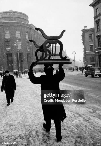 In a light snowfall, a man is carrying a Art Deco table on his head along the pavement of a street in Leningrad. Leningrad , winter 1960.