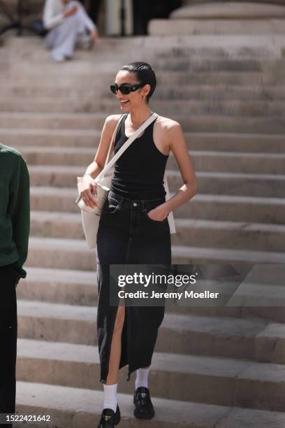 Fashion Week guest is seen wearing black sunglasses from Prada, silver earrings, a black tank top, a dark denim maxi skirt with front split, a golden...