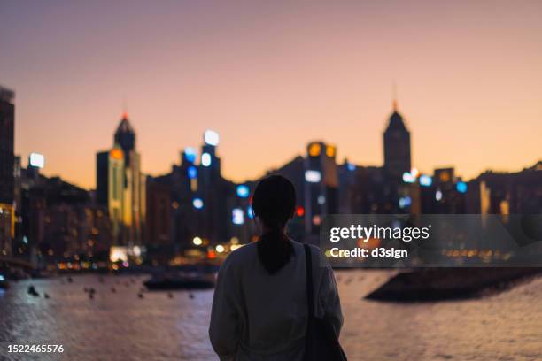 rear view of young asian female tourist overlooking the beautiful iconic city skyline of hong kong at victoria harbour at dusk. female traveller admiring the spectacular city view while exploring and strolling in the city, enjoying a vacation - hometown stockfoto's en -beelden