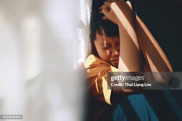 un ragazzo asiatico spaventato si copre la testa con le mani, nascondendosi nell'ombra mentre è vittima di bullismo - abuso di minore foto e immagini stock