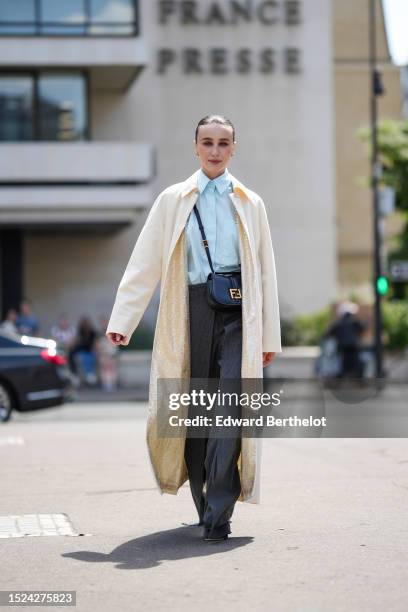Mary Leest wears gold small earrings, a pale blue shirt, a white latte long jacket, a black shiny leather with gold buckle logo crossbody bag from...
