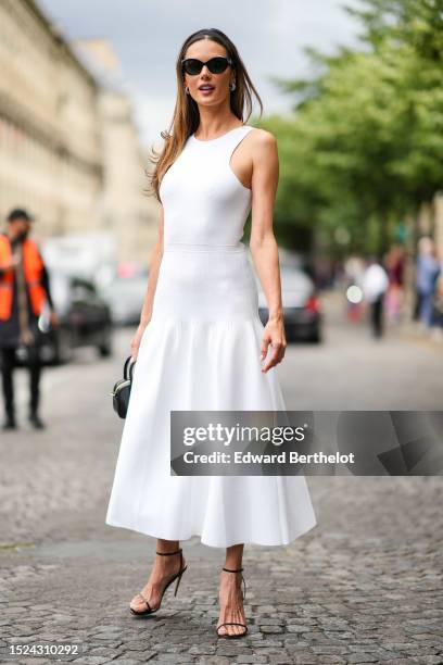 Alessandra Ambrosio wears black sunglasses, diamonds earrings, a white tank-top / embossed ribbed long dress, a black shiny varnished handbag, black...