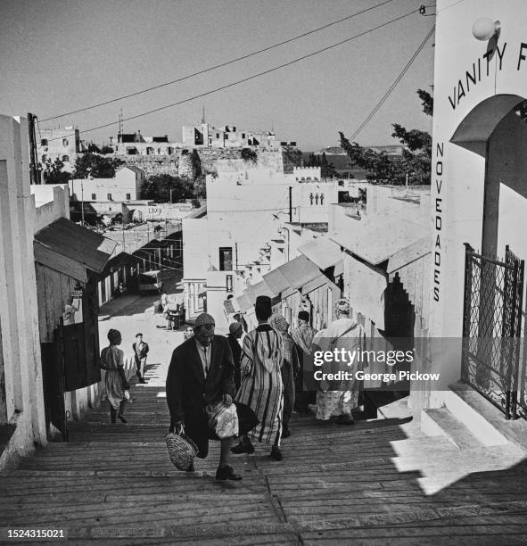 Moroccans walking on a hill with the Kasbah Palace visible in the distance, and small shops lining the slope in Tangier, Morocco, circa 1955.