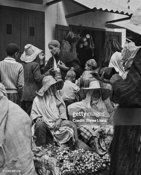 Women, both wearing floppy wide-brim straw hats, as they sit at their stall with other traders and customers in the background, in a marketplace in...