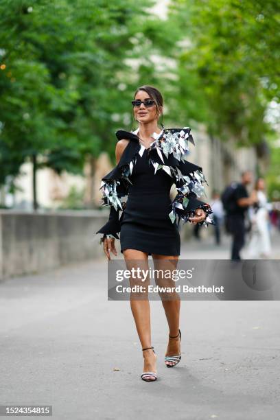 Camila Coelho wears black with rhinestones cat eyes sunglasses, silver earrings, a black cut-out shoulder / short tube dress with large embroidered...