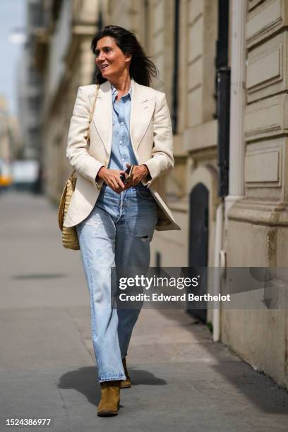 Emmanuelle Alt wears a pale blue denim shirt, a white latte blazer jacket, a beige wicker large shoulder bag, blue denim faded large pants, brown...