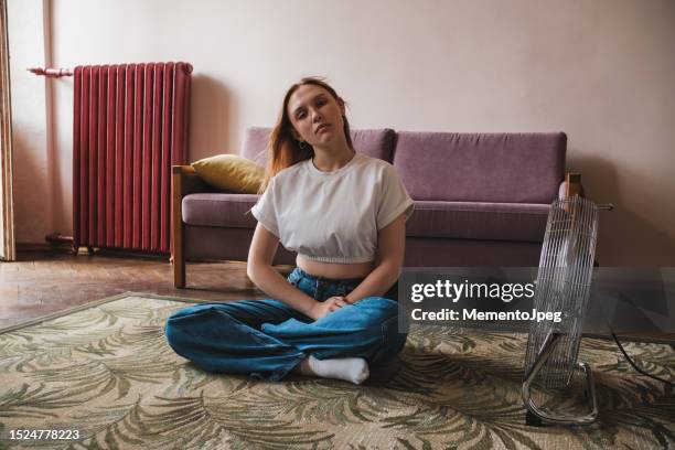 young unhappy woman suffering from summer heat at home, sitting near electric fan and looking at camera, living in apartment without air conditioning, cooling down indoors in hot weather - ac weary stock pictures, royalty-free photos & images