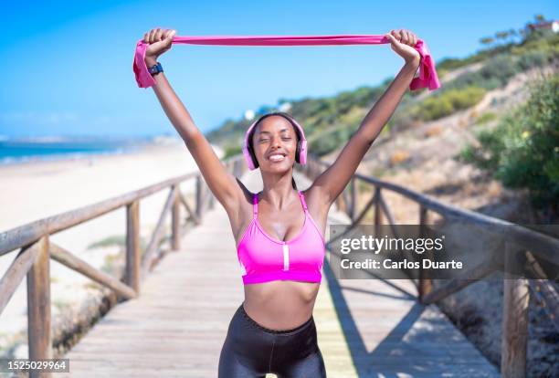 beautiful black woman exercising and listening music with her headphones in the beach in summer with resistance band around thighs - bra stock pictures, royalty-free photos & images