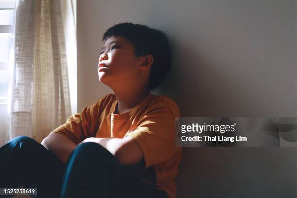 little asian boy with a sad and lonely expression hides in the shadows of a home corner. - aspergers stock pictures, royalty-free photos & images