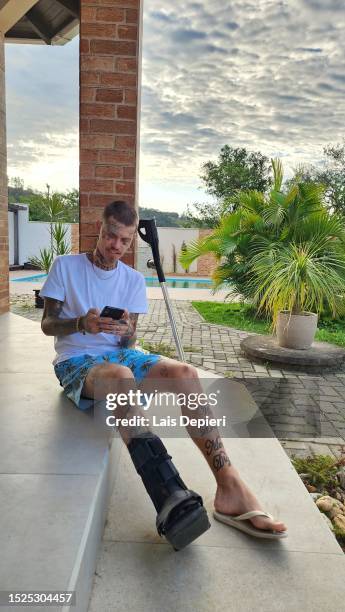 tattooed man with fracture sitting on cell phone - indian crutch stockfoto's en -beelden