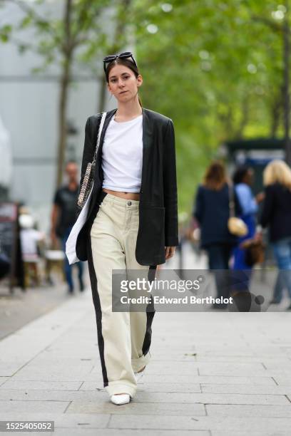 Chloe Ambre Morin wears black sunglasses, a white cropped t-shirt, a black long blazer jacket, a silver coins shoulder bag from Paco Rabanne, white...