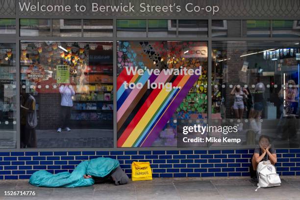 Homeless person on the pavement under a sleeping bag outside a supermarket in Soho, underneath text which reads 'We've waited too long' and beside a...