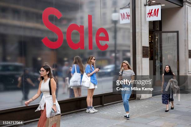 People out shopping walk past a large scale sale sign in red and silver for major high street clothing brand H&M outside their flagship store on the...