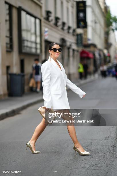 Gili Biegun wears black sunglasses, gold fringed long earrings, a white blazer jacket dress, a gold rhinestones puffy clutch, gold shiny varnished...