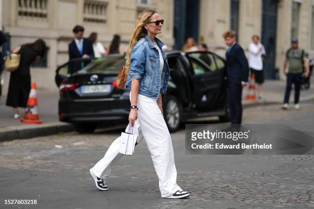 Guest wears black sunglasses, silver small earrings, a blue denim jacket, a white t-shirt, white denim large pants, a white Chanel shopping bag, a...