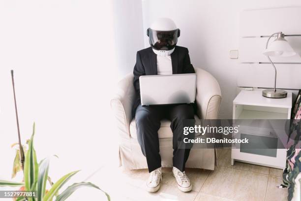 man wearing suit jacket and astronaut helmet, sitting in an armchair in a bedroom with a bay window, working with a laptop. concept of explorer, scientist, weirdness, space exploration, creativity. - space helmet stock pictures, royalty-free photos & images