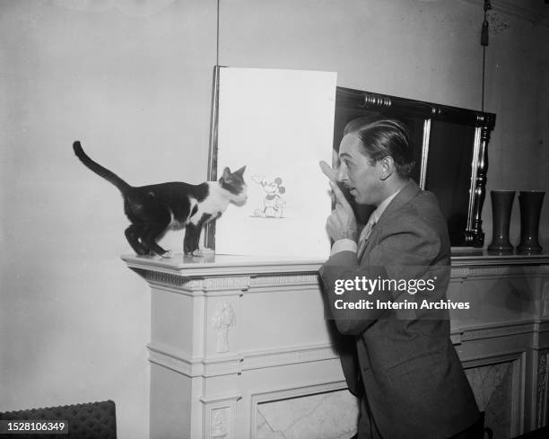 American animator and film pioneer Walt Disney holds a drawing of the cartoon character Mickey Mouse while gesturing to a cat, 1931.