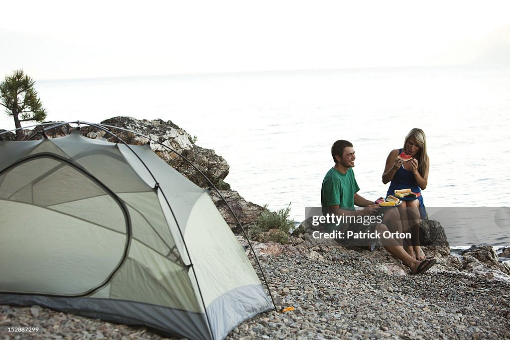 Two young adults camping smile while eating dinner next to a lake in Idaho.