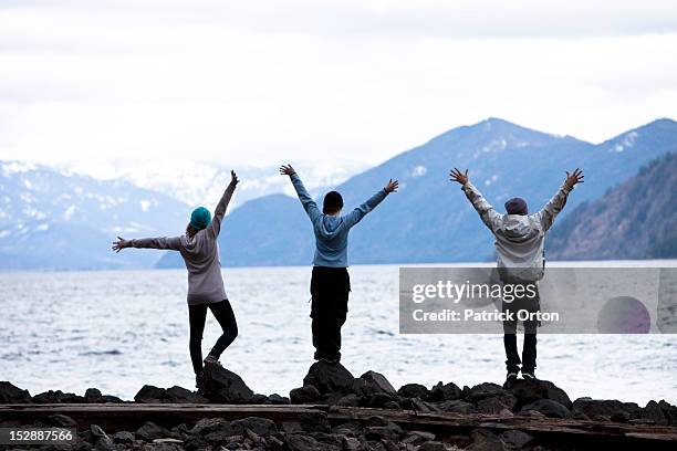 a group of three young adults standing with their arms open at the edge of a lake in idaho. - pend orielle lake stock pictures, royalty-free photos & images