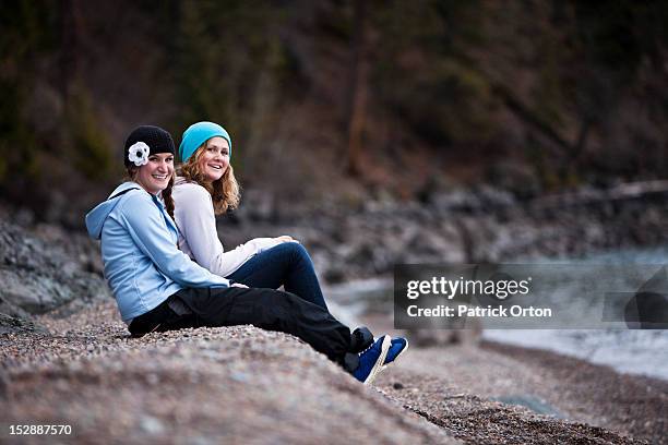 two young woman laugh and smile while sitting on a rocky beach next to a lake in idaho. - pend orielle lake stock pictures, royalty-free photos & images