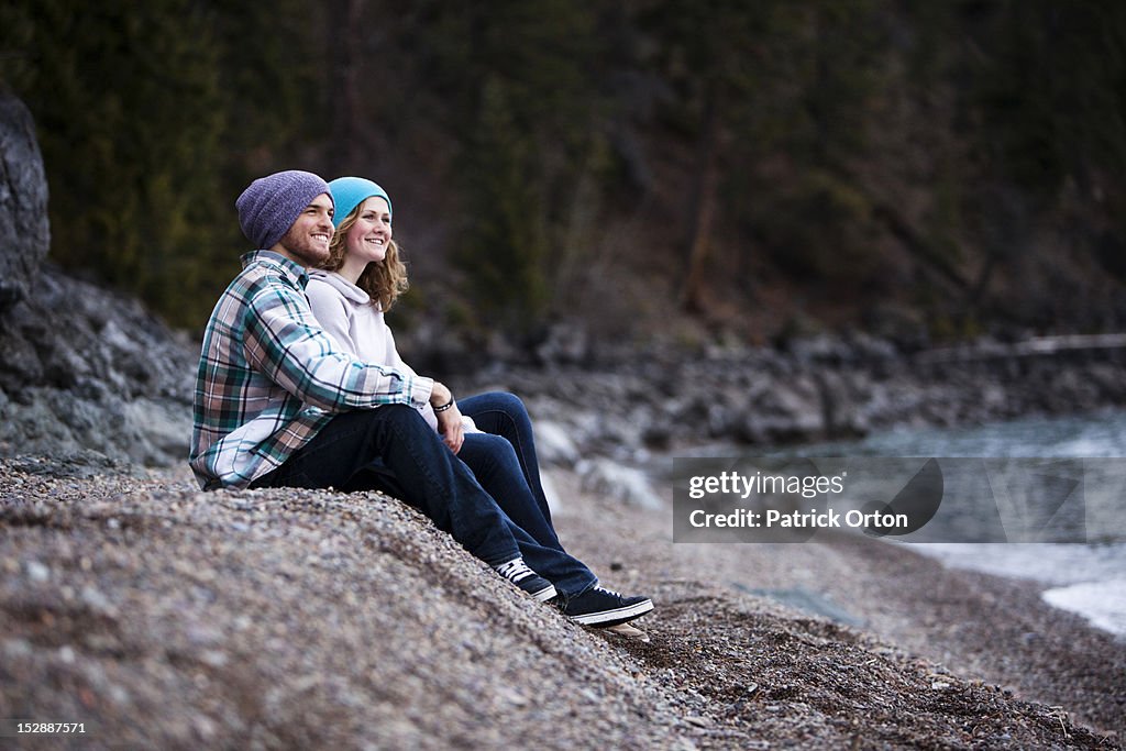 A young adult couple laugh and smile while sitting on a rocky beach next to a lake in Idaho.