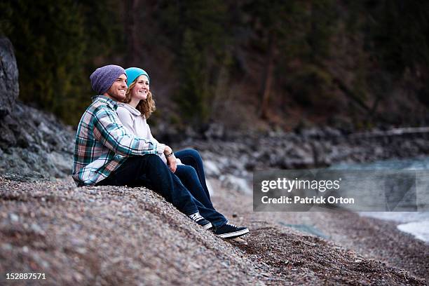 a young adult couple laugh and smile while sitting on a rocky beach next to a lake in idaho. - pend orielle lake stock pictures, royalty-free photos & images
