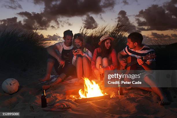 young adults sat around fire in sand dunes - feu plage photos et images de collection
