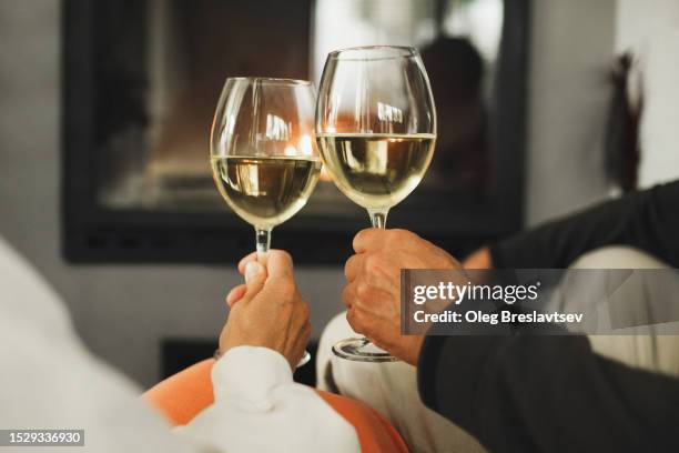 two glasses with white wine in couple's hands. romantic evening together near fireplace. - drinks stock pictures, royalty-free photos & images