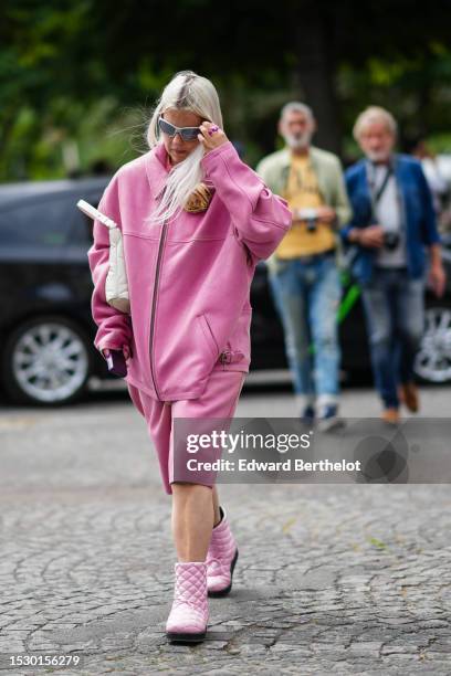 Guest wears gray sport sunglasses, a pink suede zipper oversized jacket, matching pale pink long suede shorts, a white matte leather shoulder bag,...