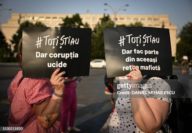 Two women cover their head from the sun with signs reading in Romanian "They all knew - But corruption kills" and "They all knew - But they're all...