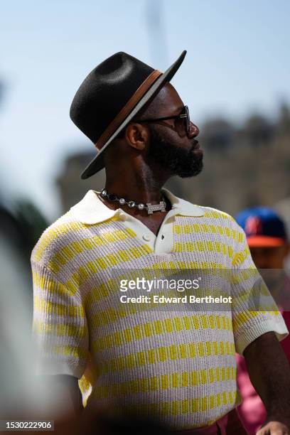 Guest wears a black felt wool with brown leather ribbon hat, black sunglasses, rhinestones and white pearls necklace, a white and yellow striped...