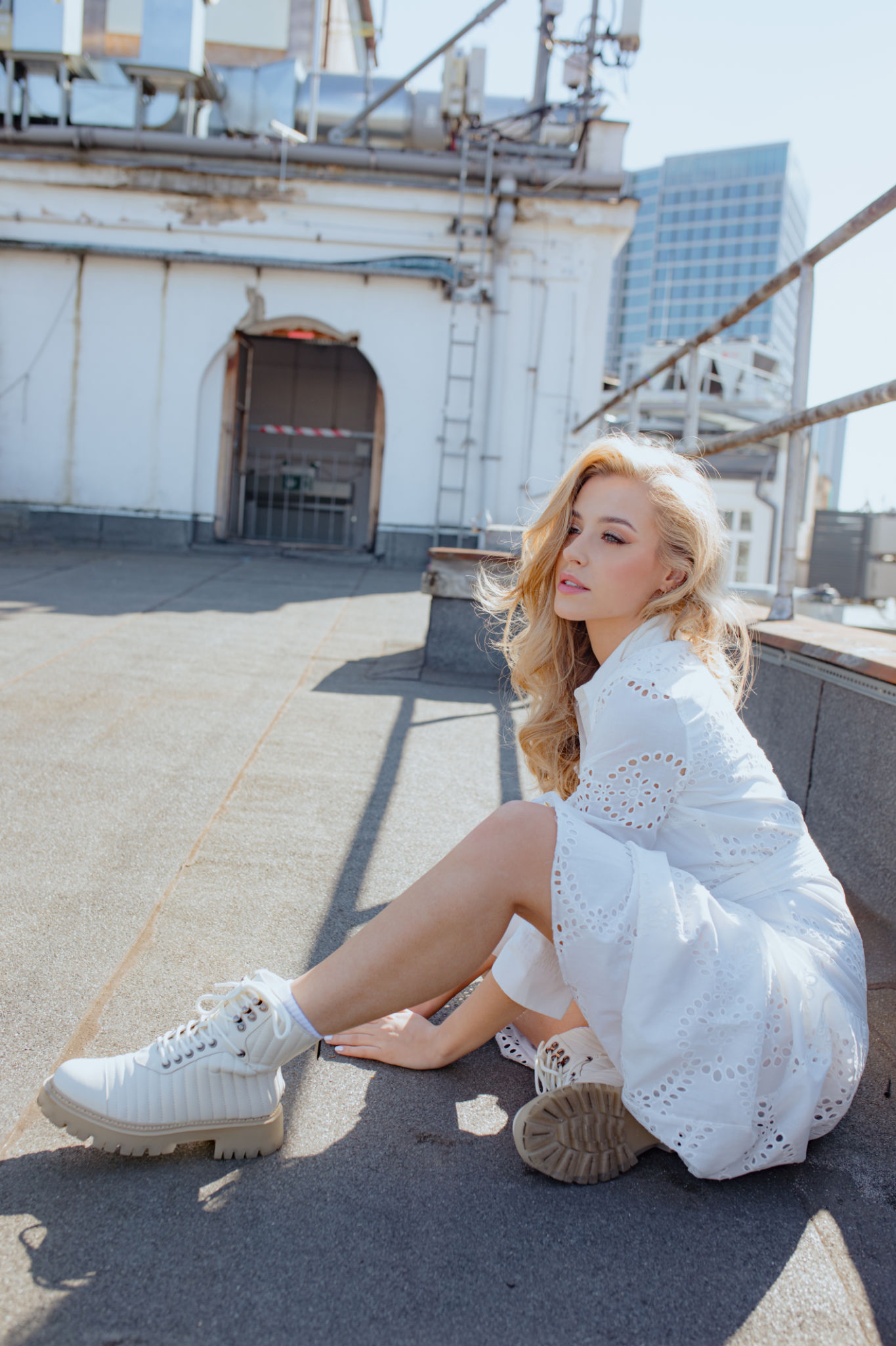 Young woman in white lace dress, laced boots sitting near fence on solar rooftop of city building and looking forward. Young woman in white lace dress, laced boots sitting near fence on solar rooftop of city building and looking forward.