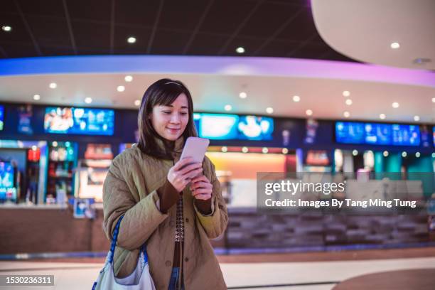 young asian woman using e-ticketing reservation on mobile app with her smartphone in front of ticket counter at cinema - taquilla lugar de comercio fotografías e imágenes de stock