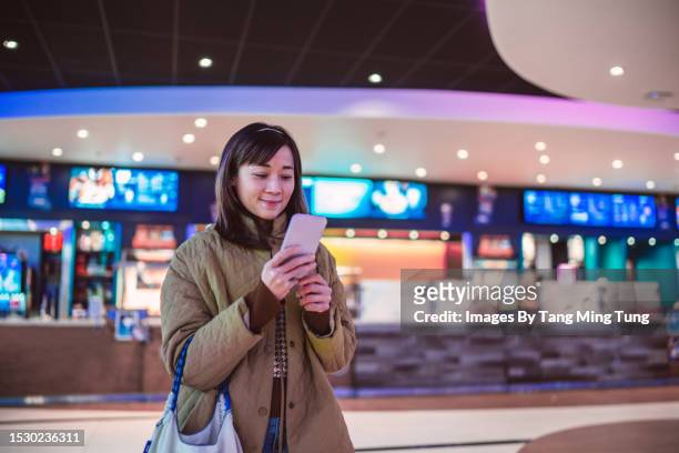 young asian woman using e-ticketing reservation on mobile app with her smartphone in front of ticket counter at cinema - balcão da bilheteira imagens e fotografias de stock