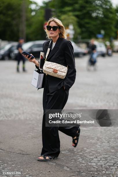 Guest wears black sunglasses, a white t-shirt, a black blazer jacket, black oversized pants, a white cut-out FF pattern crossbody bag from Fendi, a...