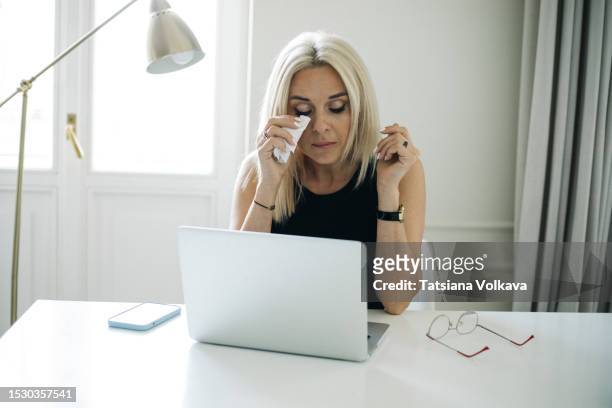 upset mature business woman sitting at table in front of laptop holding napkin to dry tears - unhappy woman blonde glasses stock pictures, royalty-free photos & images