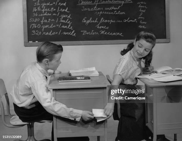 Elementary school age boy and girl sitting at their desks secretly passing a note between each other.