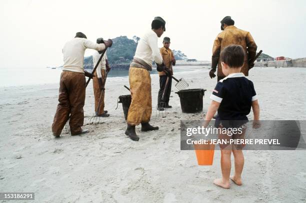 Little boy with a shovel and a bucket looks at people cleaning up a beach in Portsall, in June 1978, after the Amoco Cadiz oil spill. The Liberian...