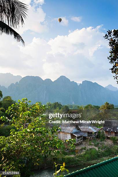 a hot air balloon hovers above a jungle village.. - vang vieng balloon stock pictures, royalty-free photos & images