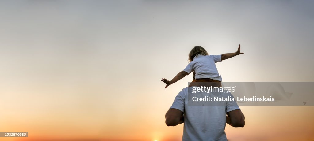 Magical Fatherhood: Enchanting Encounter of a Dad Carrying his Baby Toddler on Shoulders, Creating a World of Wonder and Joy in a Field at Sunset, Embracing the Magic of Father-Child Connections. Banner and copy space. Back view