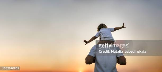magical fatherhood: enchanting encounter of a dad carrying his baby toddler on shoulders, creating a world of wonder and joy in a field at sunset, embracing the magic of father-child connections. banner and copy space. back view - father day fotografías e imágenes de stock