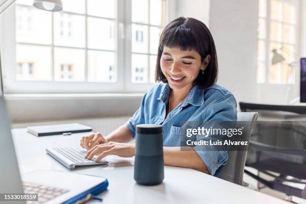 young businesswoman talking to virtual assistant speaker at her office desk - ai speaker stock-fotos und bilder