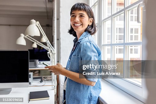 Young businesswoman standing by the window with mobile phone at startup office Young businesswoman standing by the window with mobile phone at startup office