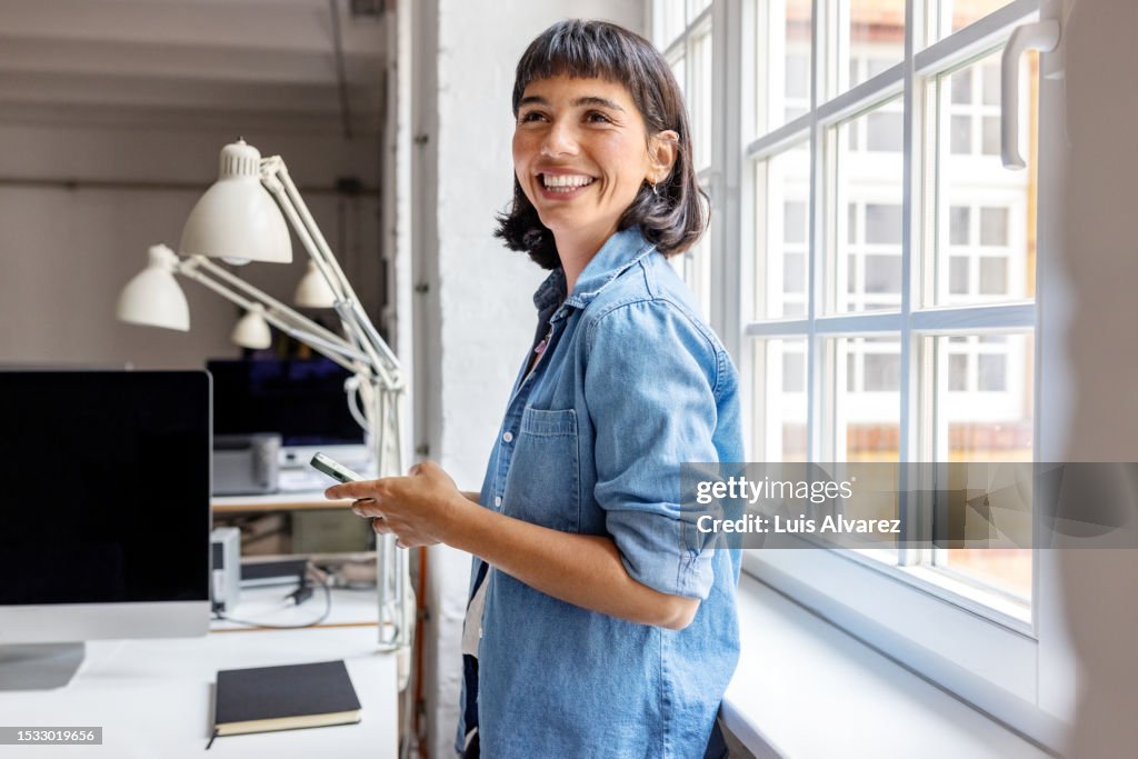 Young businesswoman standing by the window with mobile phone at startup office