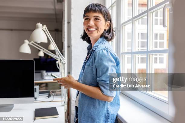 young businesswoman standing by the window with mobile phone at startup office - lifestyle imagens e fotografias de stock