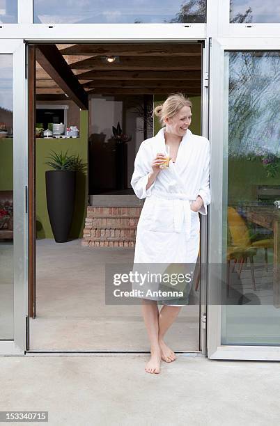 a happy woman in her bathrobe leaning in the doorway of her house - bademantel stock-fotos und bilder