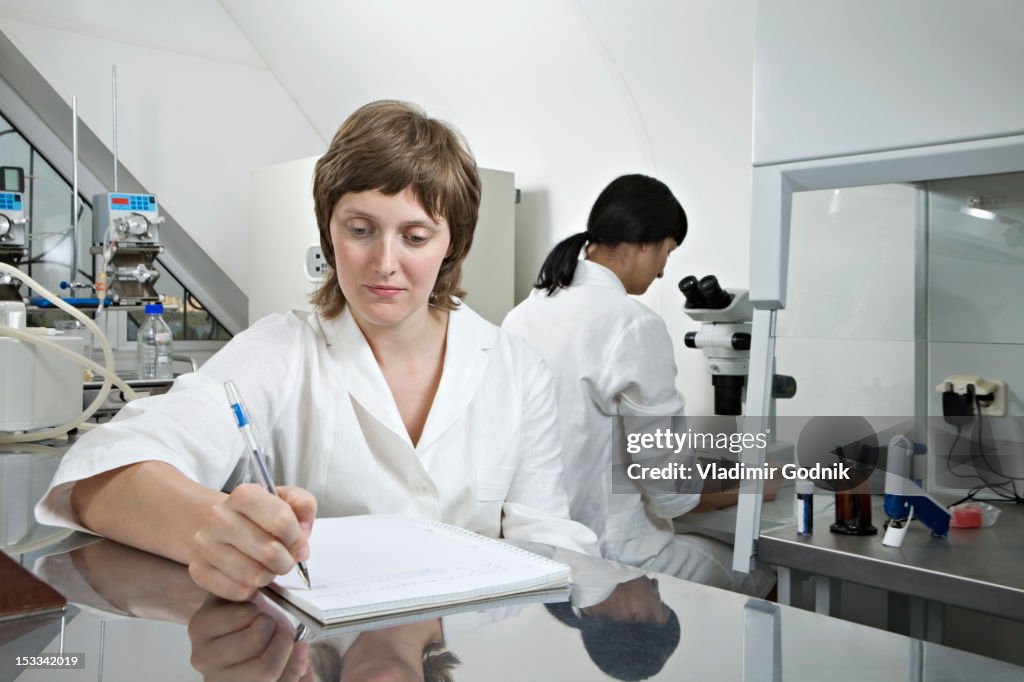 Person Writing Lab Notes A Lab Technician Writing Notes While Another Technician Works In ...